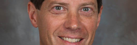 Headshot of Todd Denison wearing a black suit, blue shirt and navy blue tie on a grey background